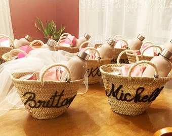 May include: A group of small woven baskets with handles, each containing a glass bottle and personalized with a name in black script. The baskets are arranged on a wooden table.