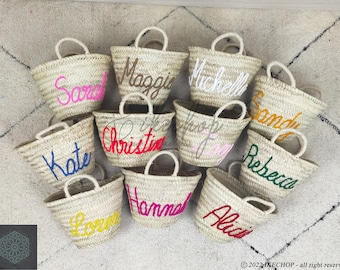 May include: A collection of 10 small, natural woven straw tote bags with colorful embroidered names, including Kate, Sarah, Maggie, Michelle, Sandy, Rebecca, Christine, Hope, Hanna, and Alicia.