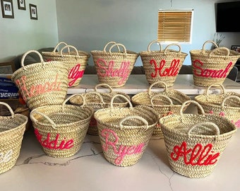 May include: A collection of nine straw tote bags with pink and red embroidered names. The bags are arranged in three rows on a white surface.