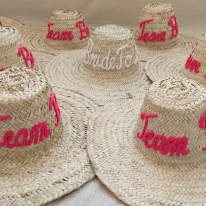 May include: A collection of woven straw sun hats. The hats have wide brims and cylindrical crowns. The crowns are embroidered with the words "Team B" in pink thread, and one hat says "Bride To Be" in white.