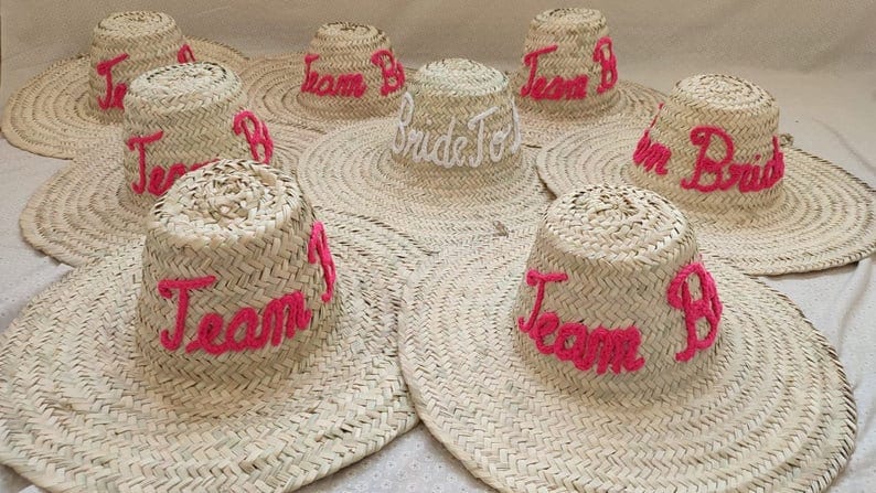 May include: A collection of woven straw sun hats. The hats have wide brims and cylindrical crowns. The crowns are embroidered with the words "Team B" in pink thread, and one hat says "Bride To Be" in white.