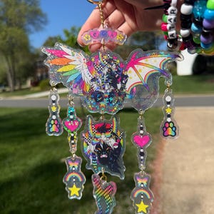 May include: A clear acrylic keychain with a gold keyring. The keychain features a large, colorful winged creature design with the word "VEGAS" at the top. Additional charms include hearts, stars, and rainbows in vibrant colors.
