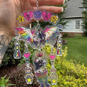 May include: A clear acrylic keychain with the word "VEGAS" spelled out in colorful letters and shapes. The keychain features a bat-like design with rainbow wings and several dangling charms with hearts, stars, and paw prints. The overall design is vibrant and eye-catching.