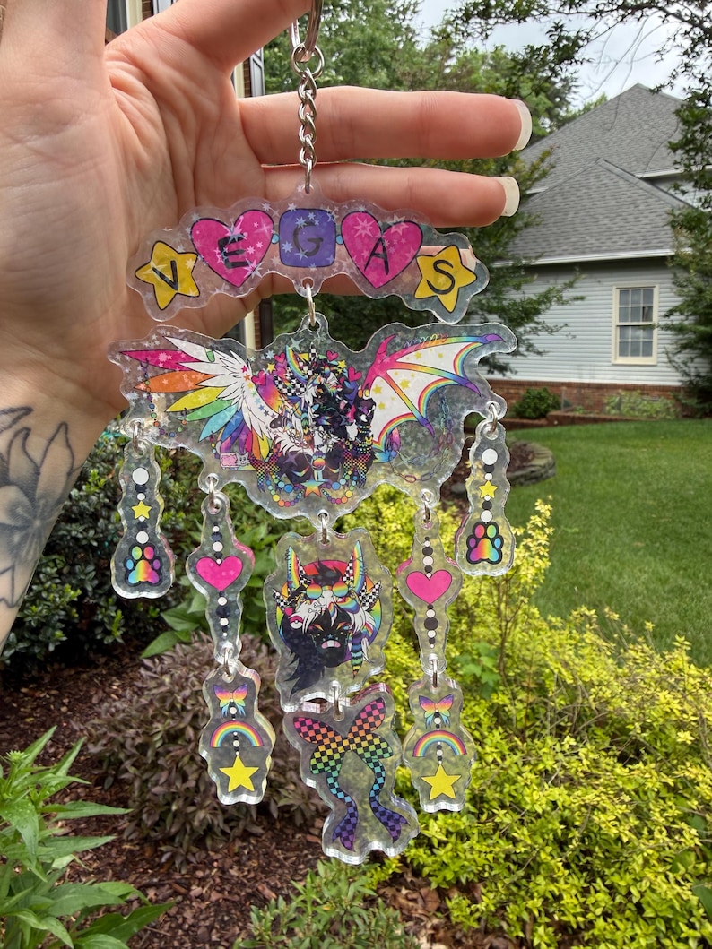 May include: A clear acrylic keychain with the word "VEGAS" spelled out in colorful letters and shapes. The keychain features a bat-like design with rainbow wings and several dangling charms with hearts, stars, and paw prints. The overall design is vibrant and eye-catching.