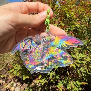 May include: A colorful, clear acrylic keychain featuring a fantastical creature with rainbow wings and intricate details. The design includes a checkered pattern and vibrant hues of pink, yellow, blue, and green, attached to a green clasp.