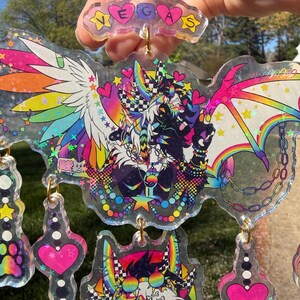 May include: A collection of colorful, transparent keychains with holographic accents. The keychains feature cartoon characters with rainbow wings, hearts, and stars. One keychain spells out "VEGAS".