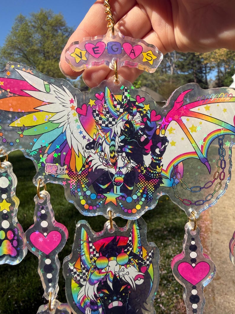May include: A collection of colorful, transparent keychains with holographic accents. The keychains feature cartoon characters with rainbow wings, hearts, and stars. One keychain spells out "VEGAS".