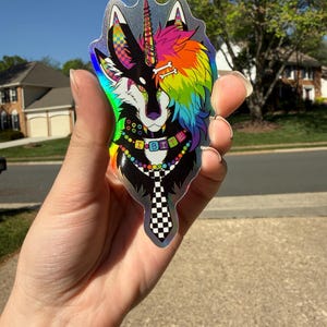 May include: Holographic sticker featuring a wolf-unicorn hybrid design. The wolf side is black and white, while the unicorn side is rainbow-colored. The sticker has a checkered tie and a collar that spells "I BITE".