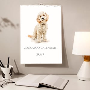 May include: A wall calendar for the year 2027 featuring a watercolour illustration of a light-coloured Cockapoo dog. The calendar is white with black text that reads COCKAPOO CALENDAR and 2027, and it is bound with a silver metal spiral wire.