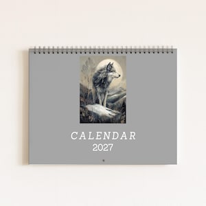 May include: A grey wall calendar for the year 2027 with a spiral binding at the top. The centre of the calendar features a rectangular illustration of a wolf standing on a rocky landscape against a large moon, with the text CALENDAR 2027 below.