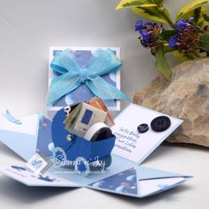 Könnte beinhalten: Eine blaue und weiße Explosionsbox mit einem blauen Kinderwagen im Inneren. Die Box ist mit Sternen und Monden verziert. Die Box ist geöffnet und der Kinderwagen ist mit Geld gefüllt. Die Box ist mit einem blauen Band gebunden. Der Text auf der Box lautet "Stemp'n'Joy" und "www.stampnjoy.handmade.de".