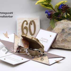 Könnte beinhalten: Eine braune und beige Explosionsbox mit einer goldenen Zahl 60 auf der Oberseite. Die Box ist geöffnet und zeigt eine kleine braune Box mit einem silbernen Armband im Inneren. Die Box ist mit einem Kartenmuster und einem goldenen Herzen verziert.
