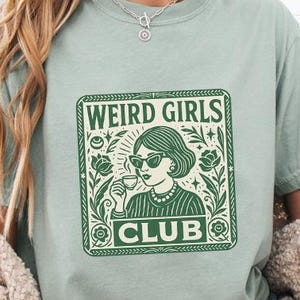 May include: A light green t-shirt with the words "WEIRD GIRLS CLUB" in a green rectangular design. The design features a woman wearing sunglasses and holding a drink. The model is wearing blue jean shorts and a cream-colored sweater.