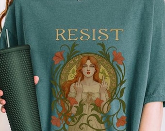 May include: Teal-colored oversized t-shirt with the word "RESIST" printed across the chest. The shirt features a central graphic of a woman with red hair, floral accents, and a defiant gesture. The shirt is made of a soft material.
