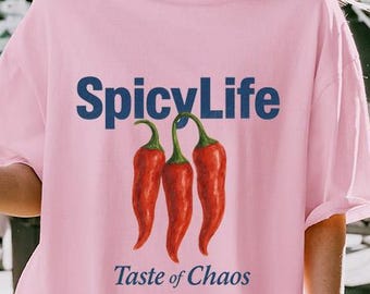 Op de afbeelding: Een lichtroze t-shirt met de tekst "SpicyLife" in marineblauw boven drie rode chilipepers. Onder de pepers staat de tekst "Taste of Chaos" in marineblauw. Het shirt heeft een casual, oversized stijl.
