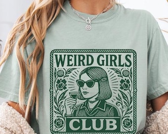 May include: A light green t-shirt with a graphic that reads "WEIRD GIRLS CLUB" in a green rectangular frame. The graphic features a woman wearing sunglasses and holding a drink. The shirt is paired with denim shorts and a cream-colored cardigan.
