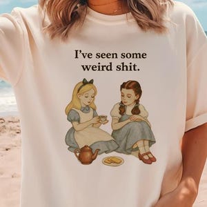 May include: Cream-colored t-shirt featuring a whimsical illustration of two girls at a tea party, with the text "I've seen some weird shit." The design includes a teapot, teacups, and cookies, creating a playful and unique aesthetic.