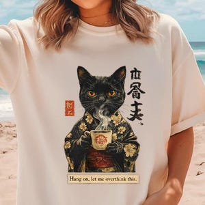 May include: Cream-colored t-shirt featuring a black cat in a kimono holding a teacup with steam rising. The shirt has Japanese text and the phrase "Hang on, let me overthink this."