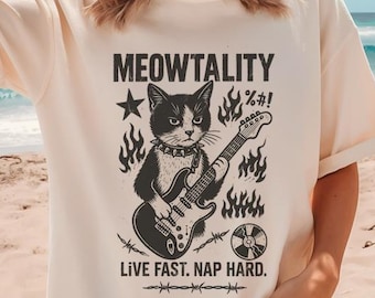 May include: Cream-colored t-shirt featuring a black and white graphic of a cat playing a guitar, with the text "MEOWTALITY" above and "LIVE FAST. NAP HARD." below. The design includes flames, a star, and a record.