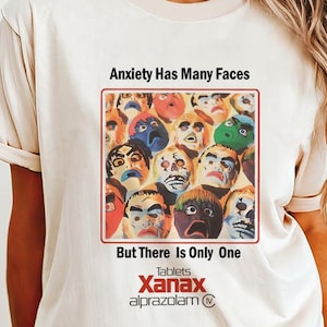 Puede incluir: Camiseta color crema con un gráfico que dice "Anxiety Has Many Faces But There Is Only One Xanax alprazolam". El gráfico muestra un collage de caras coloridas. La camiseta tiene mangas cortas y cuello redondo.