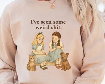 May include: A beige sweatshirt with the text "I've seen some weird shit." The graphic features two girls, a cat, and a dog. The girls are wearing blue dresses and sitting on a bench. The cat is orange and the dog is brown.