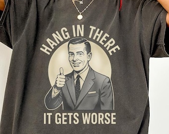 May include: Dark grey t-shirt with the text "HANG IN THERE IT GETS WORSE" and a vintage illustration of a man giving a thumbs up. The shirt has a relaxed fit and short sleeves.