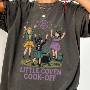 May include: Dark grey t-shirt featuring a whimsical illustration of three young witches around a cauldron. The design includes a pentagram, a tombstone, and the text "LITTLE COVEN COOK-OFF". The shirt has a vintage, slightly faded look.