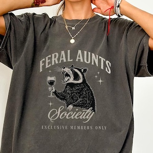 May include: Dark gray t-shirt with the text "FERAL AUNTS Society EXCLUSIVE MEMBERS ONLY". The shirt features a raccoon holding a wine glass. The raccoon is black and white with a snarling expression.