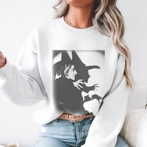 May include: White sweatshirt featuring a black and white graphic of a witch and her shadow. The design is a silhouette of a woman with a pointed hat and a shadowy figure with clawed hands. The sweatshirt is a casual, long-sleeved garment.