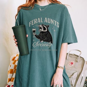 May include: Teal-colored t-shirt with the text "FERAL AUNTS Society" and a graphic of a raccoon holding a wine glass. The shirt is oversized and has a relaxed fit. A dark green tumbler is held in the left hand.