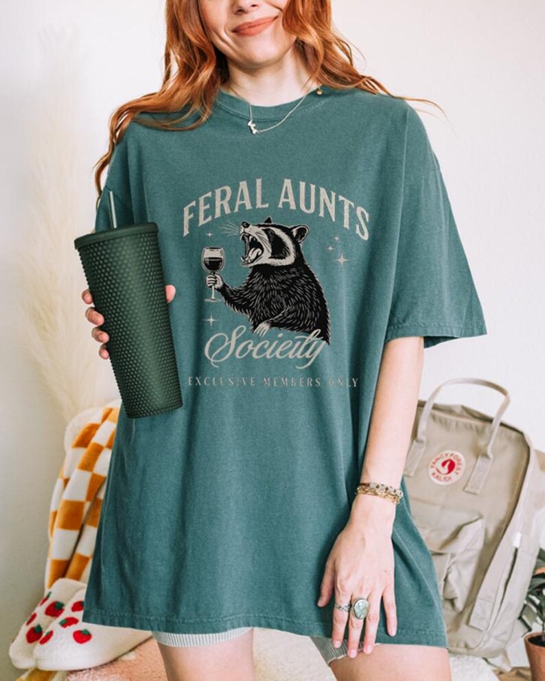 May include: Teal-colored t-shirt with the text "FERAL AUNTS Society" and a graphic of a raccoon holding a wine glass. The shirt is oversized and has a relaxed fit. A dark green tumbler is held in the left hand.