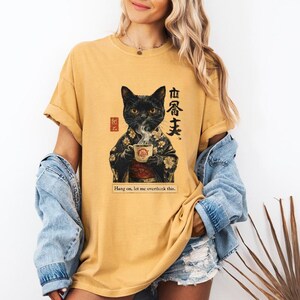May include: Mustard yellow t-shirt featuring a black cat in a kimono holding a teacup. The graphic includes Japanese text and the phrase "Hang on, let me overthink this."