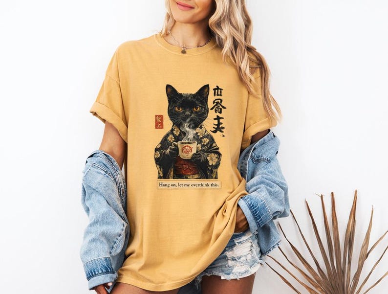 May include: Mustard yellow t-shirt featuring a black cat in a kimono holding a teacup. The graphic includes Japanese text and the phrase "Hang on, let me overthink this."