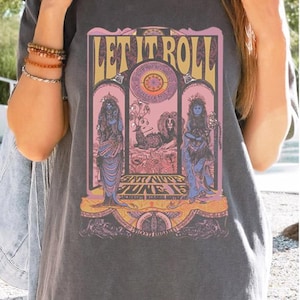 May include: Dark gray t-shirt with a colorful graphic design. The design features the text "LET IT ROLL" above an illustration of figures and a lion. The shirt also has the text "SAT NITE JUNE 15".