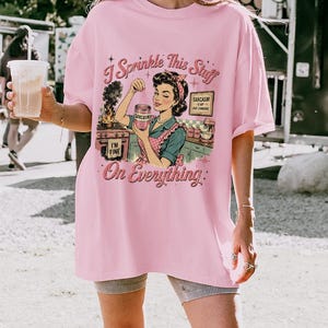 May include: A light pink t-shirt with a retro graphic of a woman in a kitchen, holding a jar labeled "Sarcasm." Text on the shirt reads "I Sprinkle This Stuff On Everything." The woman is wearing a red and white polka dot apron.