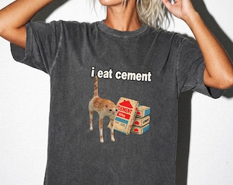 May include: A charcoal grey short-sleeve t-shirt featuring a graphic of an orange cat standing next to a stack of cement bags. The text "i eat cement" is printed in white letters above the cat. The shirt has a faded, washed-out appearance.