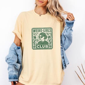 May include: A light yellow t-shirt with a green graphic that reads "WEIRD GIRLS CLUB." The graphic features a woman wearing sunglasses and holding a drink. The shirt is a relaxed fit.