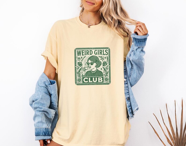 May include: A light yellow t-shirt with a green graphic that reads "WEIRD GIRLS CLUB." The graphic features a woman wearing sunglasses and holding a drink. The shirt is a relaxed fit.