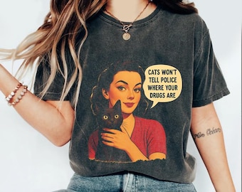 May include: Dark gray t-shirt featuring a retro illustration of a woman holding a black cat. The image includes the text "Cats won't tell police where your drugs are" in a speech bubble. The shirt is a vintage style.