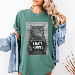 May include: A green t-shirt featuring a black and white image of a cat holding a sign that reads "I HATE PEOPLE." The cat is depicted as if it were a mugshot, with a cigarette in its mouth.
