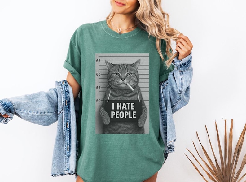May include: A green t-shirt featuring a black and white image of a cat holding a sign that reads "I HATE PEOPLE." The cat is depicted as if it were a mugshot, with a cigarette in its mouth.