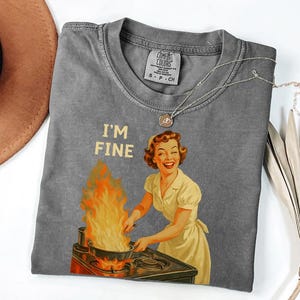 May include: A gray Comfort Colors t-shirt featuring a retro illustration of a woman smiling while cooking with flames. The text "I'M FINE" is above the illustration. A silver necklace with a pendant is on the shirt.