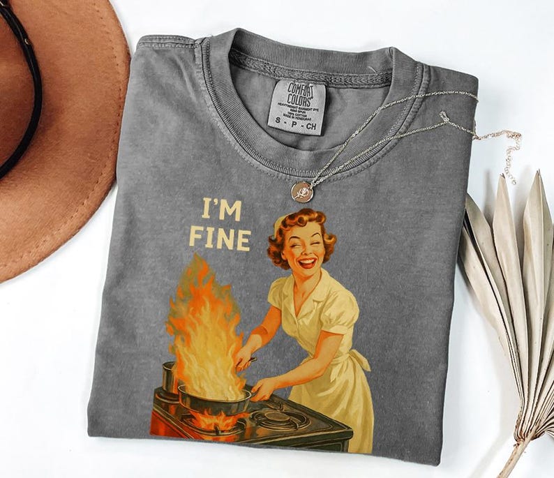 May include: A gray Comfort Colors t-shirt featuring a retro illustration of a woman smiling while cooking with flames. The text "I'M FINE" is above the illustration. A silver necklace with a pendant is on the shirt.