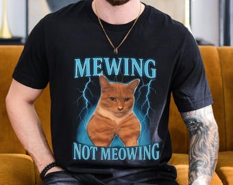 Könnte beinhalten: Schwarzes T-Shirt mit einer Grafik einer orangefarbenen Katze und dem Text "MEWING NOT MEOWING" in Hellblau. Das Katzenbild wird von blauen Blitzen umrahmt.