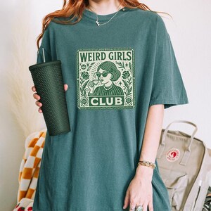May include: Teal-colored t-shirt with a green graphic that reads "WEIRD GIRLS CLUB." The graphic features a woman with sunglasses and a drink, surrounded by floral elements. The shirt is a relaxed fit.
