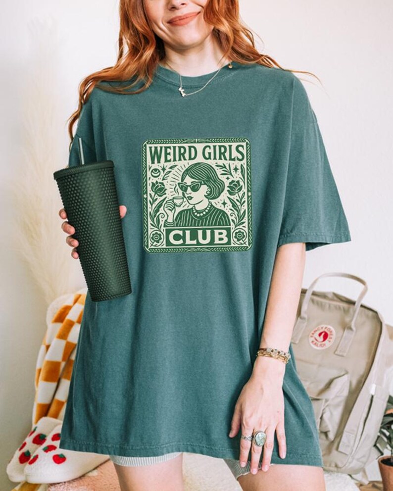May include: Teal-colored t-shirt with a green graphic that reads "WEIRD GIRLS CLUB." The graphic features a woman with sunglasses and a drink, surrounded by floral elements. The shirt is a relaxed fit.