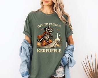 May include: Olive green t-shirt with a graphic of a raccoon wearing a cowboy hat and cape riding a snail. The text "OFF TO CAUSE A KERFUFFLE" is printed on the shirt in a serif font. The shirt has short sleeves and a relaxed fit.
