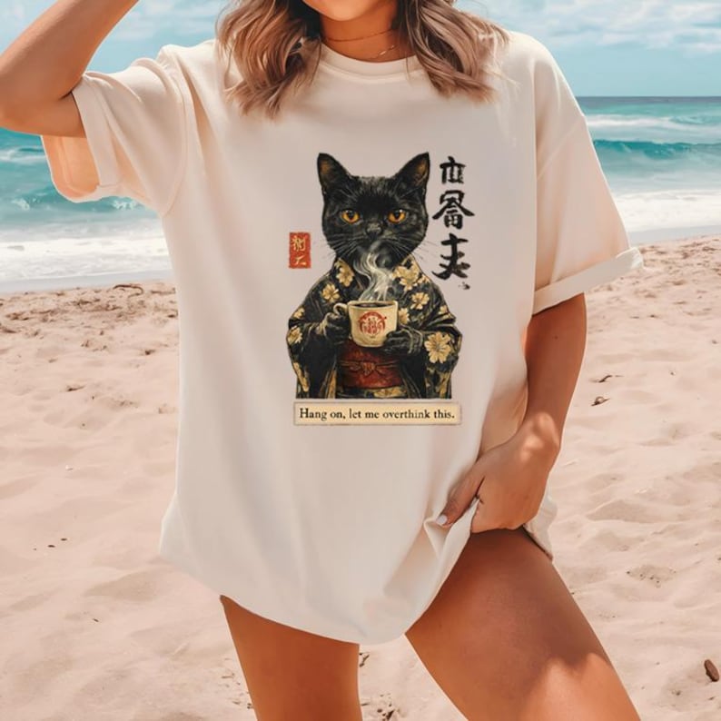 May include: Cream-colored t-shirt featuring a black cat in a kimono holding a teacup with steam rising. The shirt has Japanese text and the phrase "Hang on, let me overthink this."