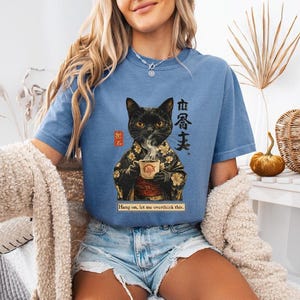 May include: A blue t-shirt featuring a black cat in a kimono holding a teacup. The graphic includes Japanese text and the phrase "Hang on, let me overthink this." The shirt is a relaxed fit.