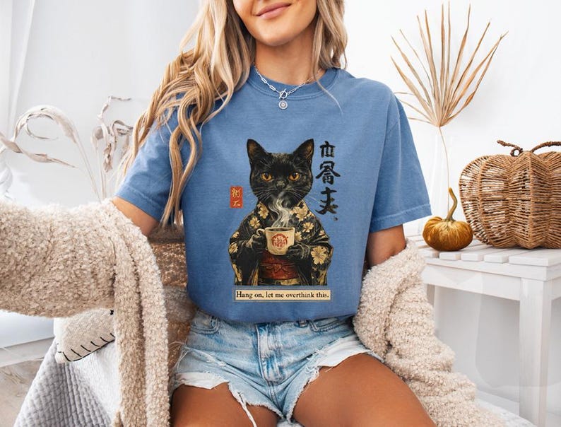 May include: A blue t-shirt featuring a black cat in a kimono holding a teacup. The graphic includes Japanese text and the phrase "Hang on, let me overthink this." The shirt is a relaxed fit.